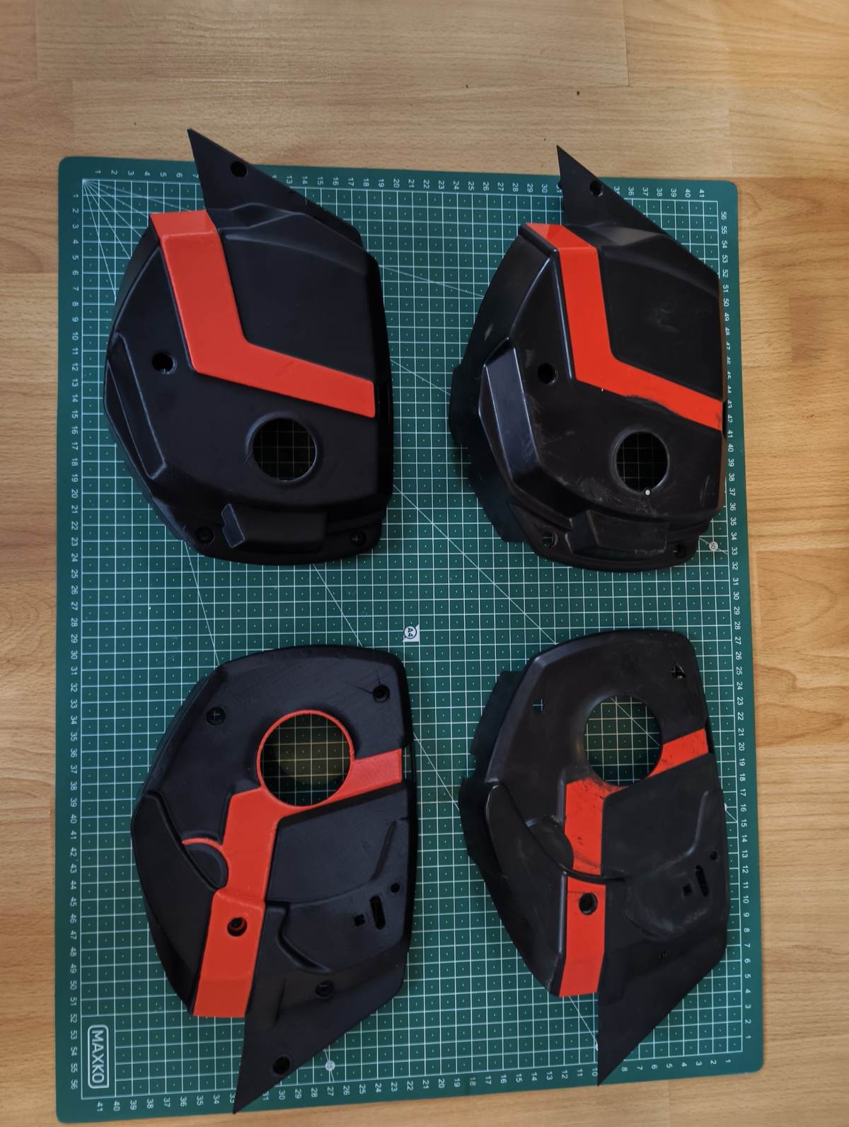 Vier schwarze Gehäuseabdeckungen mit roten Streifen auf einer grünen Schneidematte mit Zentimeterraster. Links zwei saubere Nachbauten, rechts die abgenutzten Originale mit Kratzern.